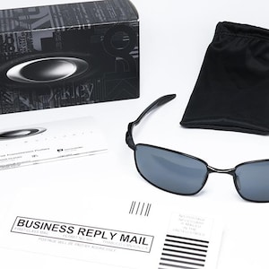Puede incluir: Gafas de sol rectangulares negras con lentes grises, una funda de transporte negra y una caja negra con el logotipo de Oakley. Las gafas de sol van acompa&ntilde;adas de una tarjeta blanca y un sobre de respuesta comercial.