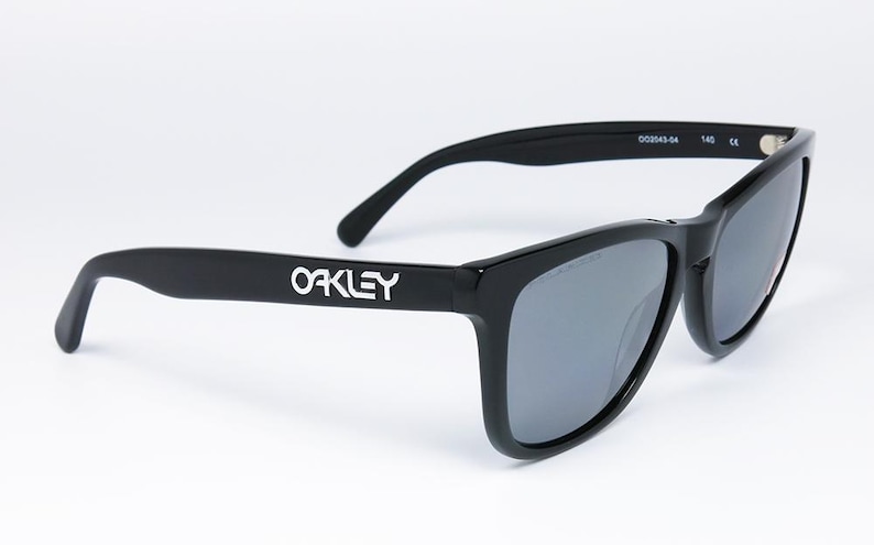 May include: Black Oakley sunglasses with gray lenses. The Oakley logo is displayed in white on the left arm. The sunglasses have a classic, rectangular frame design and are suitable for outdoor activities.