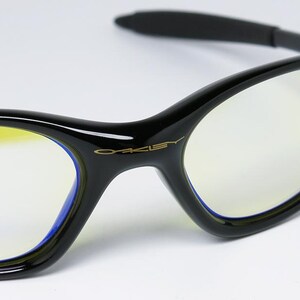 Peut inclure: Lunettes &agrave; monture noire avec des verres teint&eacute;s de jaune. Les lunettes ont un design &eacute;l&eacute;gant et incurv&eacute; avec le nom de la marque "Oakley" en lettres dor&eacute;es sur la monture. Les verres sont bord&eacute;s d'une bordure bleue.
