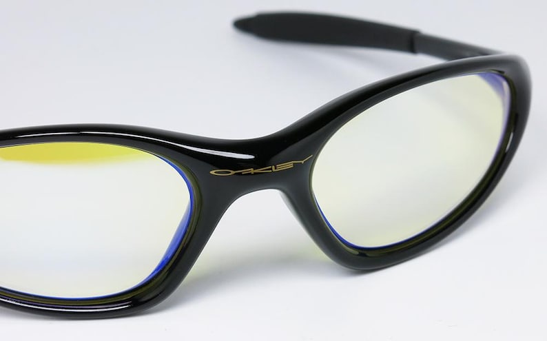 Peut inclure: Lunettes &agrave; monture noire avec des verres teint&eacute;s de jaune. Les lunettes ont un design &eacute;l&eacute;gant et incurv&eacute; avec le nom de la marque "Oakley" en lettres dor&eacute;es sur la monture. Les verres sont bord&eacute;s d'une bordure bleue.