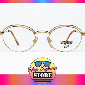 Può includere: Occhiali rotondi dorati con un design a doppio bordo. Gli occhiali hanno lenti trasparenti e il marchio "MOSCHINO by Persol" è stampato sulla lente destra.