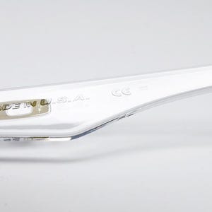 以下が含まれることがあります： 透明なメガネのテンプルのクローズアップ。 「MADE IN U.S.A.」の文字と数字「03-247 58 16」が見えます。 テンプルには、ヒンジの近くに紫色のアクセントがあります。
