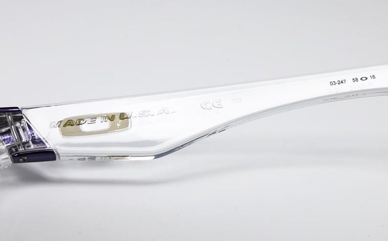 以下が含まれることがあります： 透明なメガネのテンプルのクローズアップ。 「MADE IN U.S.A.」の文字と数字「03-247 58 16」が見えます。 テンプルには、ヒンジの近くに紫色のアクセントがあります。