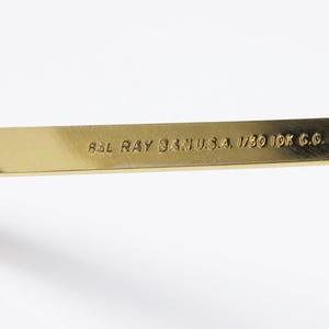 Puede incluir: Primer plano de una patilla de gafas dorada con el texto "B&L RAY BAN U.S.A. 1/30 10K G.O." La bisagra es visible a la izquierda. La patilla es de color oro met&aacute;lico brillante.