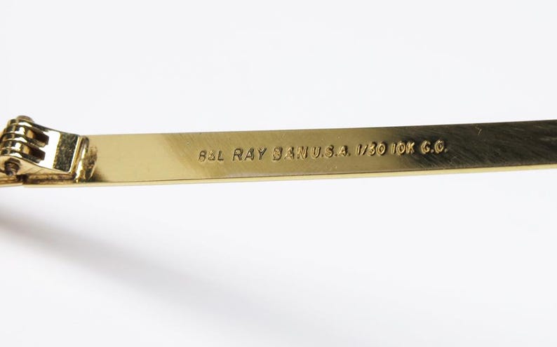 Puede incluir: Primer plano de una patilla de gafas dorada con el texto "B&L RAY BAN U.S.A. 1/30 10K G.O." La bisagra es visible a la izquierda. La patilla es de color oro met&aacute;lico brillante.