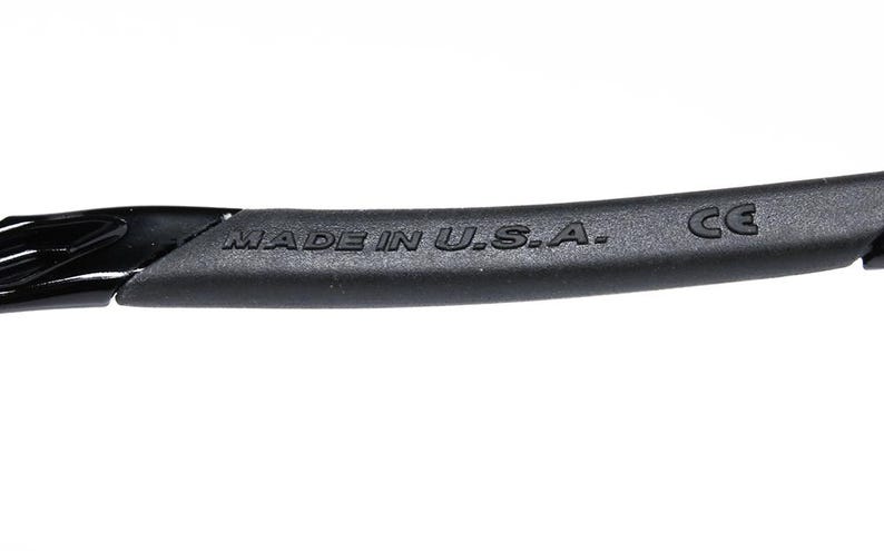 May include: Close-up of a black eyewear frame with the text "MADE IN U.S.A." and the CE mark. The frame is made of a matte black material and has a curved design. The image is set against a white background.