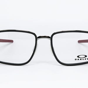 May include: Black-framed eyeglasses with a square shape and clear lenses. The temples have a red accent. The Oakley logo is visible on the lower right lens. The glasses are set against a white background.
