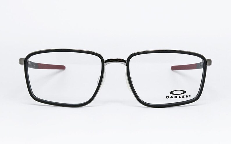 May include: Black-framed eyeglasses with a square shape and clear lenses. The temples have a red accent. The Oakley logo is visible on the lower right lens. The glasses are set against a white background.