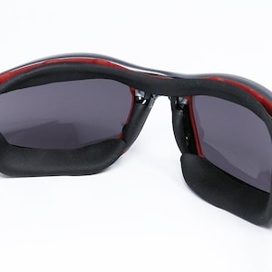 Puede incluir: Un par de gafas de sol negras con detalles rojos y lentes grises. Las gafas tienen un dise&ntilde;o deportivo con una montura flexible y un puente nasal acolchado.