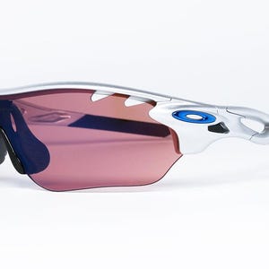 May include: Silver framed sunglasses with a pink lens and blue accents. The sunglasses have a wraparound design and a black temple.