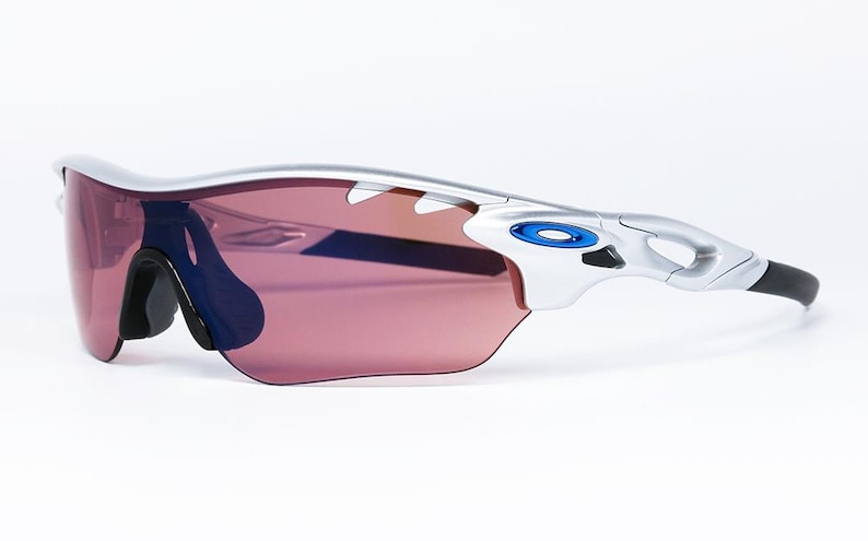 May include: Silver framed sunglasses with a pink lens and blue accents. The sunglasses have a wraparound design and a black temple.