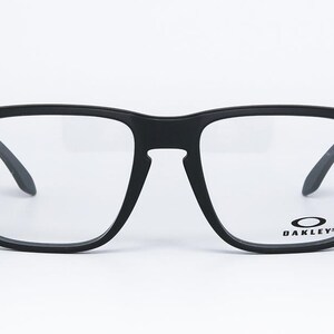 May include: Black square-framed spectacles with clear lenses. The glasses have a matte finish and the Oakley logo is visible on the lower right lens. The temples are dark and angled. The frame has small silver accents on the corners.