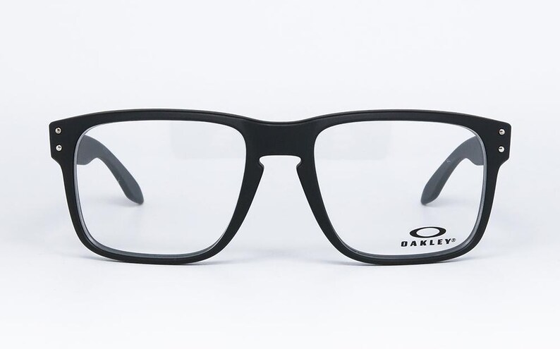 May include: Black square-framed spectacles with clear lenses. The glasses have a matte finish and the Oakley logo is visible on the lower right lens. The temples are dark and angled. The frame has small silver accents on the corners.