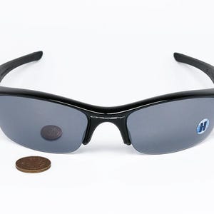 K&ouml;nnte beinhalten: Schwarze Sonnenbrille mit grauen Gl&auml;sern. Die Sonnenbrille hat ein halb rahmenloses Design und einen geschwungenen Rahmen. Eine M&uuml;nze dient zum Gr&ouml;&szlig;envergleich. Die Gl&auml;ser haben einen kleinen Aufkleber.