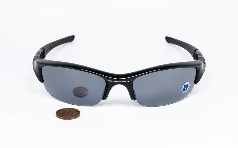 K&ouml;nnte beinhalten: Schwarze Sonnenbrille mit grauen Gl&auml;sern. Die Sonnenbrille hat ein halb rahmenloses Design und einen geschwungenen Rahmen. Eine M&uuml;nze dient zum Gr&ouml;&szlig;envergleich. Die Gl&auml;ser haben einen kleinen Aufkleber.