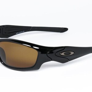 May include: Black sunglasses with brown lenses. The sunglasses have a sleek, curved design with a glossy finish. The arms of the sunglasses are slightly curved and feature a small logo near the hinges. The lenses are a warm brown colour.