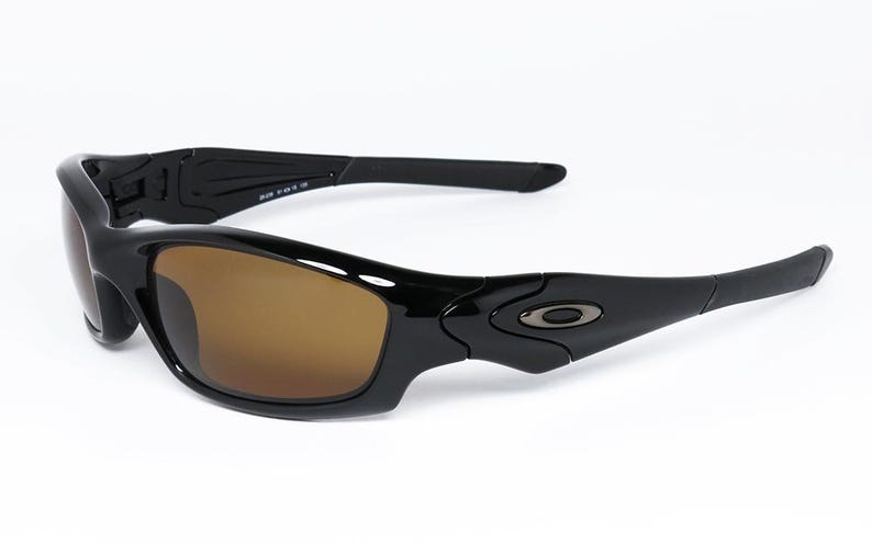 May include: Black sunglasses with brown lenses. The sunglasses have a sleek, curved design with a glossy finish. The arms of the sunglasses are slightly curved and feature a small logo near the hinges. The lenses are a warm brown colour.