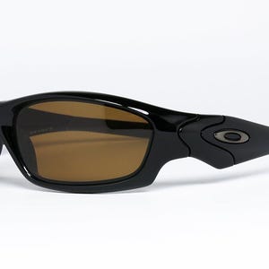 May include: Black sunglasses with brown lenses. The sunglasses have a curved, sporty design with a glossy finish. The arms feature a subtle logo detail. The lenses are a warm brown colour.