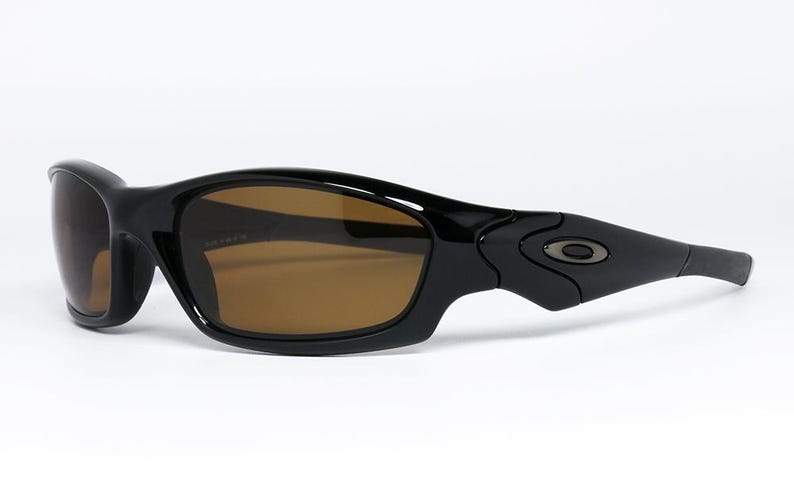 May include: Black sunglasses with brown lenses. The sunglasses have a curved, sporty design with a glossy finish. The arms feature a subtle logo detail. The lenses are a warm brown colour.