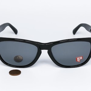 May include: Black sunglasses with gray lenses. The sunglasses have a classic design with a thick frame. A penny is placed near the sunglasses for size comparison. A red and white logo is visible on the right lens.