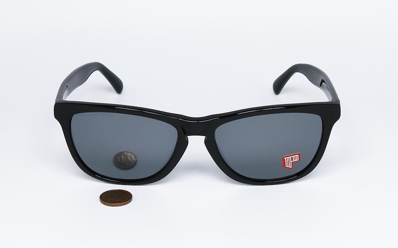 May include: Black sunglasses with gray lenses. The sunglasses have a classic design with a thick frame. A penny is placed near the sunglasses for size comparison. A red and white logo is visible on the right lens.