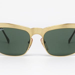 Puede incluir: Gafas de sol doradas con lentes verde oscuro. Las gafas de sol tienen un dise&ntilde;o de cejas distintivo y el logotipo de Ray-Ban es visible en la lente izquierda. La montura tiene un acabado de metal cepillado.