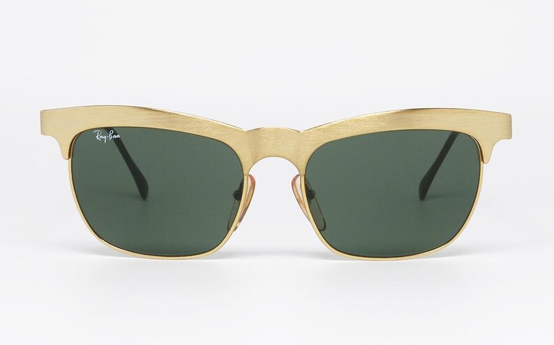 Puede incluir: Gafas de sol doradas con lentes verde oscuro. Las gafas de sol tienen un dise&ntilde;o de cejas distintivo y el logotipo de Ray-Ban es visible en la lente izquierda. La montura tiene un acabado de metal cepillado.