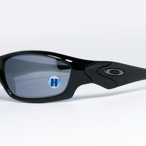 May include: Black sunglasses with gray lenses. The sunglasses have a curved frame and a small logo on the temple. A blue and white sticker with the letter "H" is on the lens.