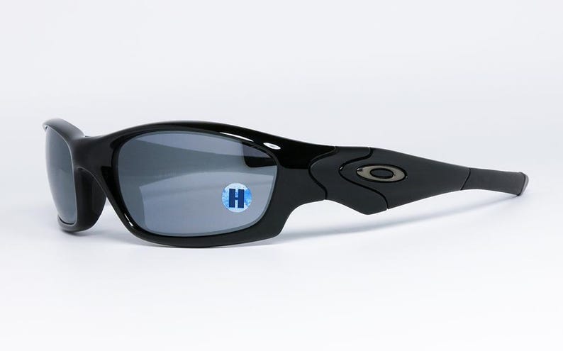 May include: Black sunglasses with gray lenses. The sunglasses have a curved frame and a small logo on the temple. A blue and white sticker with the letter "H" is on the lens.