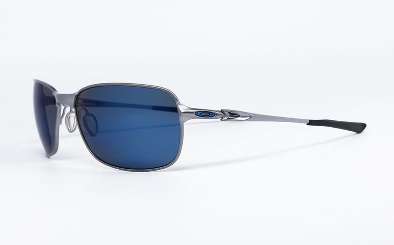 May include: Gray metal sunglasses with blue lenses. The sunglasses have a rectangular frame and a sleek, modern design. The temples are thin and silver, with black ear pieces. The lenses are a deep blue color.