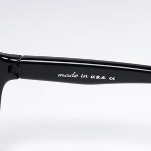 Puede incluir: Primer plano de gafas de sol negras con lentes grises. La montura es gruesa y brillante. El texto "made in U.S.A. ce" est&aacute; impreso en blanco en la varilla.