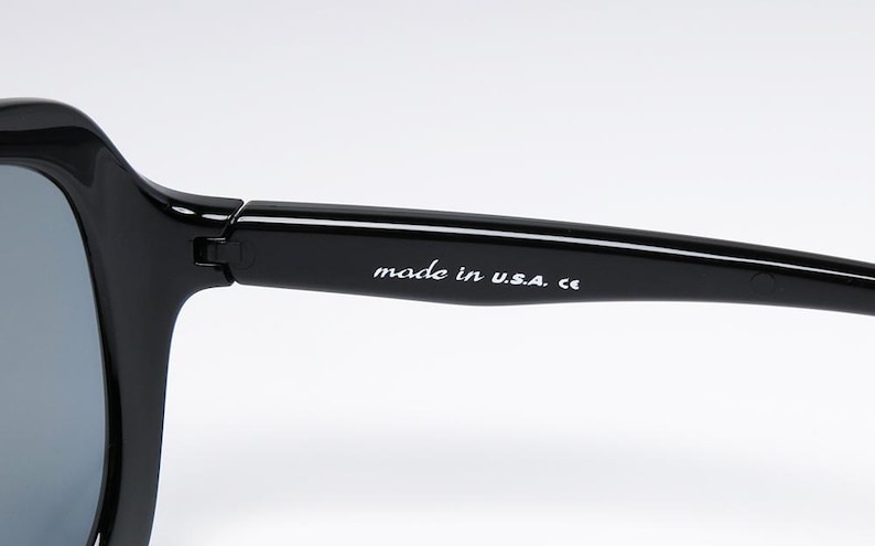 Puede incluir: Primer plano de gafas de sol negras con lentes grises. La montura es gruesa y brillante. El texto "made in U.S.A. ce" est&aacute; impreso en blanco en la varilla.