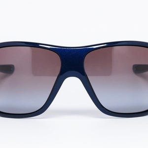 May include: Dark blue sunglasses with a gradient lens. The sunglasses have a thick frame and a rounded shape. The lenses transition from a dark brown at the top to a lighter blue at the bottom.