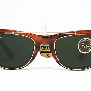 May include: Brown tortoiseshell Ray-Ban sunglasses with green lenses and gold-colored metal accents. The sunglasses have a clip-on design and a small tag with text. The Ray-Ban logo is visible on the frame.