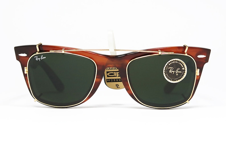 May include: Brown tortoiseshell Ray-Ban sunglasses with green lenses and gold-colored metal accents. The sunglasses have a clip-on design and a small tag with text. The Ray-Ban logo is visible on the frame.