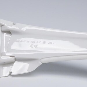 May include: Close-up of white sunglasses with dark lenses. The frame is glossy and features the text "MADE IN U.S.A." and the CE mark. The design is sleek and modern.