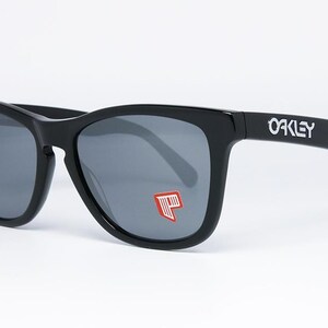 May include: Black Oakley sunglasses with gray lenses. The Oakley logo is visible on the temple. A red and white graphic is on the left lens. The sunglasses have a classic, square frame.