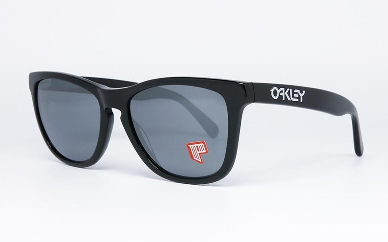 May include: Black Oakley sunglasses with gray lenses. The Oakley logo is visible on the temple. A red and white graphic is on the left lens. The sunglasses have a classic, square frame.