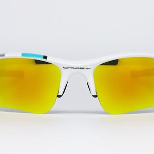 May include: White sports sunglasses with yellow-orange mirrored lenses. The sunglasses have a white frame with black accents and a small teal stripe on the top. The Oakley logo is visible on the bridge.