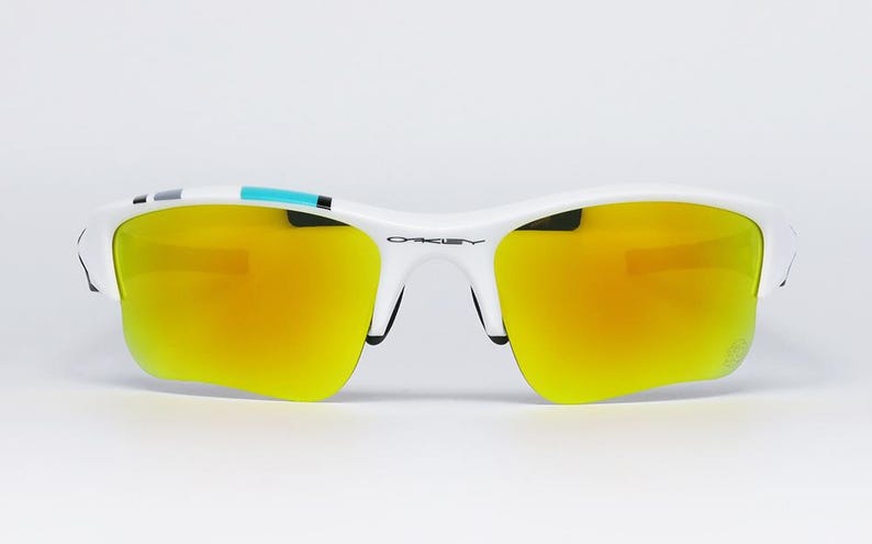 May include: White sports sunglasses with yellow-orange mirrored lenses. The sunglasses have a white frame with black accents and a small teal stripe on the top. The Oakley logo is visible on the bridge.
