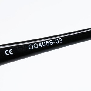 Puede incluir: Primer plano de una patilla de gafas negra con el texto "CE" y "OO4059-03" impreso en blanco. La patilla es brillante y parece estar hecha de pl&aacute;stico o un material similar. El fondo es blanco liso.