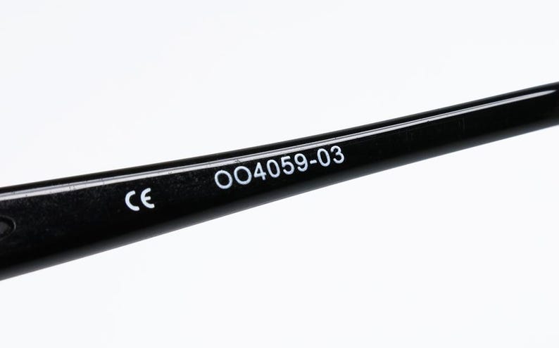Puede incluir: Primer plano de una patilla de gafas negra con el texto "CE" y "OO4059-03" impreso en blanco. La patilla es brillante y parece estar hecha de pl&aacute;stico o un material similar. El fondo es blanco liso.
