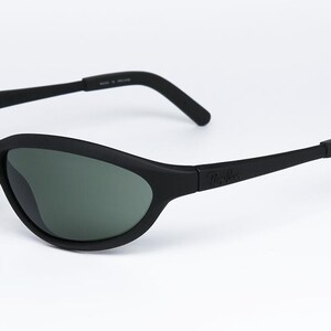 Pode incluir: &Oacute;culos de sol pretos com acabamento fosco. As lentes s&atilde;o de cor verde escuro. A arma&ccedil;&atilde;o tem um design curvo e o nome da marca &eacute; vis&iacute;vel na haste.