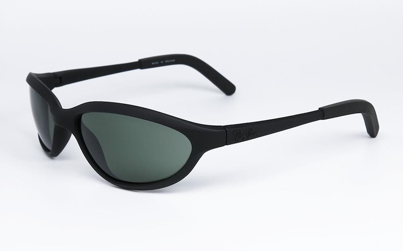 Pode incluir: &Oacute;culos de sol pretos com acabamento fosco. As lentes s&atilde;o de cor verde escuro. A arma&ccedil;&atilde;o tem um design curvo e o nome da marca &eacute; vis&iacute;vel na haste.