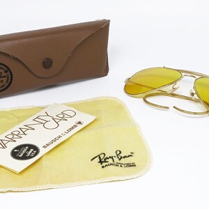 K&ouml;nnte beinhalten: Eine goldene Aviator-Sonnenbrille mit gelben Gl&auml;sern. Die Sonnenbrille ist ge&ouml;ffnet und liegt auf einer wei&szlig;en Oberfl&auml;che. Ein braunes Lederetui und ein gelbes Reinigungstuch mit dem Ray-Ban-Logo sind ebenfalls sichtbar.