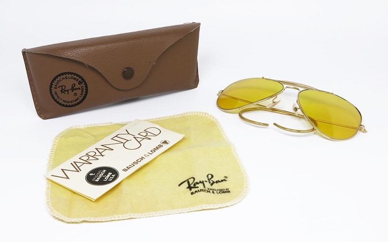 K&ouml;nnte beinhalten: Eine goldene Aviator-Sonnenbrille mit gelben Gl&auml;sern. Die Sonnenbrille ist ge&ouml;ffnet und liegt auf einer wei&szlig;en Oberfl&auml;che. Ein braunes Lederetui und ein gelbes Reinigungstuch mit dem Ray-Ban-Logo sind ebenfalls sichtbar.