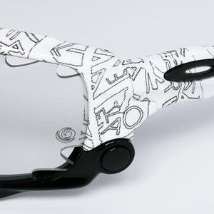 May include: Close-up of a pair of sunglasses with a white frame featuring a black graffiti-style design. The lenses are clear, and the arms are black. The Oakley logo is visible on the frame.