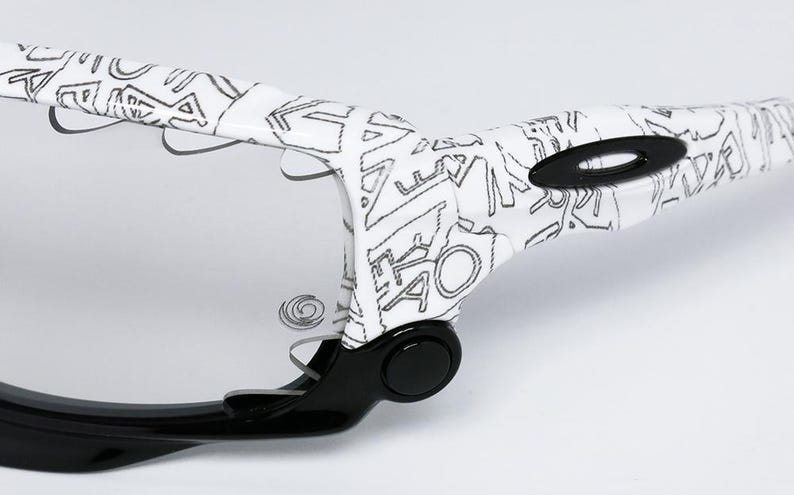 May include: Close-up of a pair of sunglasses with a white frame featuring a black graffiti-style design. The lenses are clear, and the arms are black. The Oakley logo is visible on the frame.