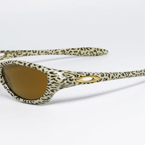 May include: Leopard print sunglasses with brown lenses. The frames and arms feature a cream and black spotted pattern. The sunglasses have a gold-colored logo on the temples. The lenses are a warm brown color.