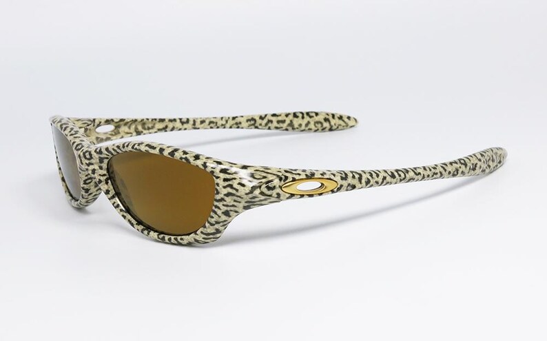 May include: Leopard print sunglasses with brown lenses. The frames and arms feature a cream and black spotted pattern. The sunglasses have a gold-colored logo on the temples. The lenses are a warm brown color.